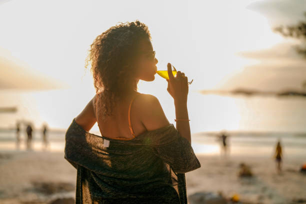 열대 해변에 좌초할 때 칵테일 주스를 마시는 아프리카 여성. 노란 비키니를 입고 자연의 아름다움을 만끽하는 젊은 여행자. 방랑벽과 여행 개념. - luxury women chaise longue fashion 뉴스 사진 이미지