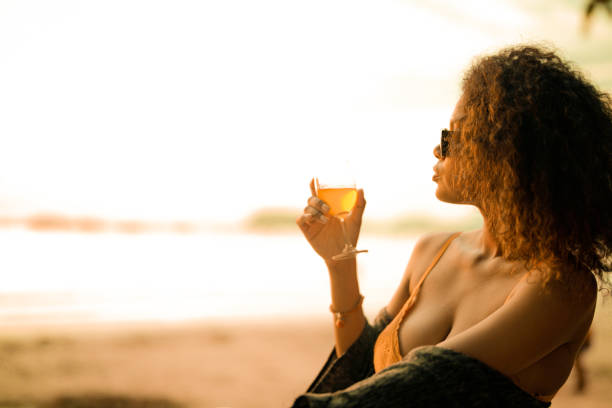 열대 해변의 벤치 해변에 앉아 칵테일 주스를 마시는 아프리카 여성. 노란 비키니를 입고 자연의 아름다움을 차갑게 하는 젊은 여행자. 방랑벽과 여행 개념. - luxury women chaise longue fashion 뉴스 사진 이미지
