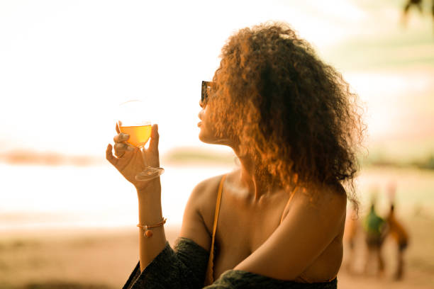 열대 해변의 벤치 해변에 앉아 칵테일 주스를 마시는 아프리카 여성. 노란 비키니를 입고 자연의 아름다움을 차갑게 하는 젊은 여행자. 방랑벽과 여행 개념. - luxury women chaise longue fashion 뉴스 사진 이미지