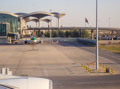 An Iraqi Airways Boeing B737 MAX 8 plane, registration YI-ASX, parked at Queen Alia International Airport near Amman. She will depart in 20 minutes as flight IA164 to Baghdad.  The overpass provides an exit from dropping passengers off to the departure area of the terminal: on the other side (not visible) is the arrival overpass.  A car park is visible under the overpass.  This is relatively expensive and consequently, many people park their car on the side of the main access road and come to collect their relatives or passenger only after they pass through immigration and customs, incurring a smaller fee to enter and pickup.  This image was taken from the upper deck of a departing Emirates A380-842 before sunset on 4 May 2023.
