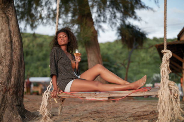 열대 해변에서 그네에 앉아 칵테일 주스를 마시는 아프리카 여자. 노란 비키니를 입고 자연의 아름다움을 차갑게 하는 젊은 여행자. 방랑벽과 여행 개념. - luxury women chaise longue fashion 뉴스 사진 이미지