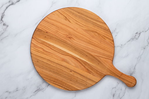Round empty wooden charcuterie serving board with handle on a white or gray marble table, top view. Kitchenware, template with copy space. Flat lay design, mockup. Layout, cooking concept, frame.