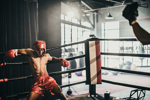 Photo of a boxer training with a punching bag in a boxing hall. There is a boxing ring behind him.