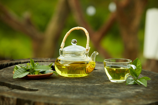 A tea cup and a teapot full of tea inside with a dish of green tea leaves placed on a wooden pedestal. Green tea can help you sleep well, anti-aging, lose weight