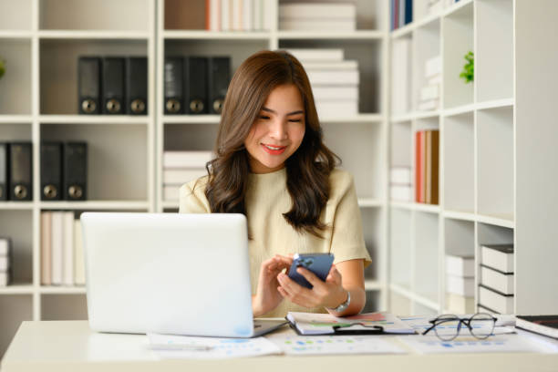 istock Satisfied asian female financial advisor chatting with client online on smart phone and using laptop at workstation. 1487907342