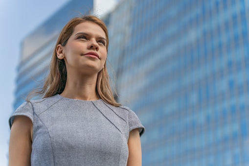 Low angle portrait shot of a confident and successful young businesswoman in the city with modern corporate skyscrapers in background. Female leadership. Determination to success. Businesswoman on the move. Connecting business relationship across the city.