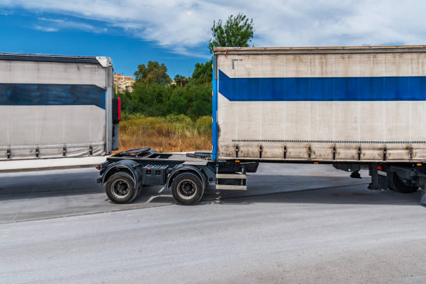 Back view of two dump trucks drives out of town on a flat asphalt road among fields against a blue sky with clouds on a sunny day. Road works on an intercity highway