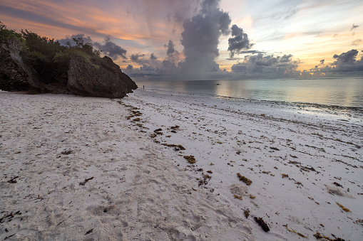 This breathtaking photo captures the beauty of a sunrise on Diani Beach in Kenya. The vibrant orange and yellow hues of the sun's rays illuminating the sky and water create a stunning contrast against the serene, turquoise ocean. In the distance, massive rainy clouds loom over the Indian Ocean, adding a sense of depth and drama to the scene. It's a moment of natural beauty that is both awe-inspiring and humbling