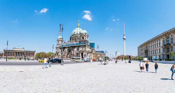 This panoramic photo captures the bustling pedestrian way along Karl-Liebknecht-Straße on a sunny cloudless day in Berlin. On the left side of the street stands the impressive Altes Museum, while the iconic Berliner Dom cathedral and the Fernsehturm tower dominate the center of the composition. In the distance, St. Marienkirche can be seen towering over the city skyline.