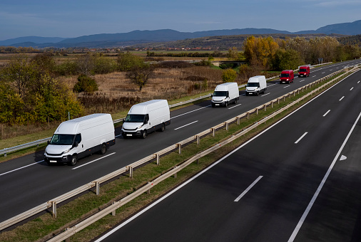 White and red minivans are moving in a convoy on the highway. White and red delivery van on the highway. White and red modern delivery small shipment cargo courier van moving fast on motorway road to city urban suburb.