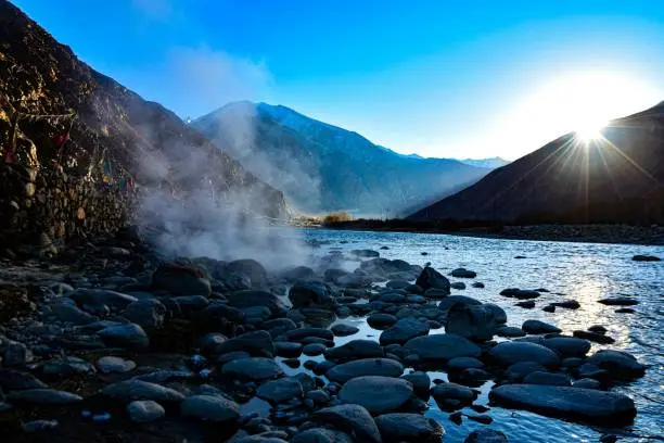 The sun rises peeking from the mountain as the river shines and steam from natural hotspring is seen.