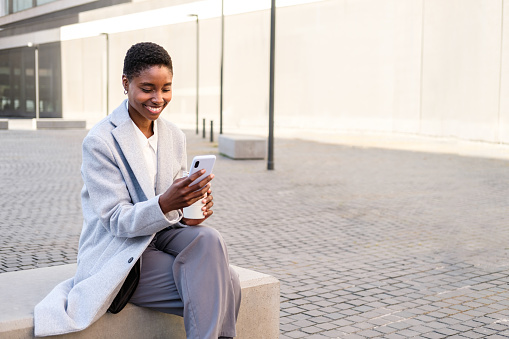 horizontal portrait of an elegant young african american woman. She is sitting near office building and using her smart phone