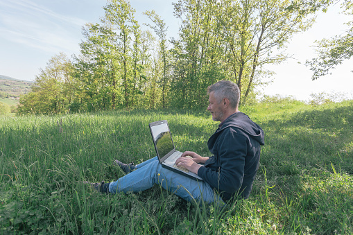 Adult man is using laptop for his work outdoors in nature on sunny springtime day. Note:  on laptop screen is reflection of background.