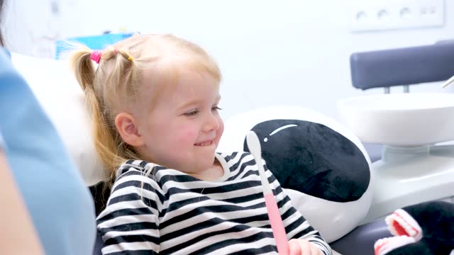 little girl in hands with toothbrush smiles bit lip and waves head negative sign no she sits in dental chair afraid she is worried brush teeth correctly first visit to dentist pediatric dentistry