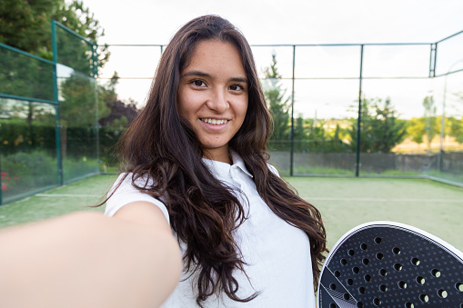 Young smiling woman taking a selfie with smart phone on padel court with racket