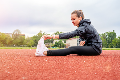 Young female athlete runner sitting on running track and stretching her legs, warming up.