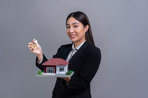 Confident Asian businesswoman holding house model, advertising home loan with smile. Real estate agent with sample house model in hand on isolated background for business advertisements. Jubilant
