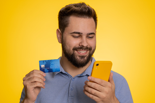 Sincere caucasian man customer using credit bank card and smartphone while transferring money, purchases online shopping, payment. Finance and internet. Happy guy isolated alone on yellow background