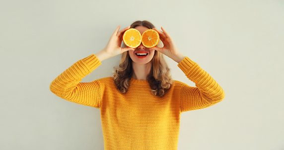 Summer portrait of happy cheerful smiling woman covering her eyes with slices of orange and looking for something on gray background