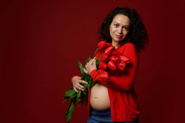 multiethnische schöne schwangere frau mit einem niedlichen tulpenstrauß, die mit nacktem bauch auf isoliertem rotem hintergrund posiert. - mothers day mother single flower family stock-fotos und bilder