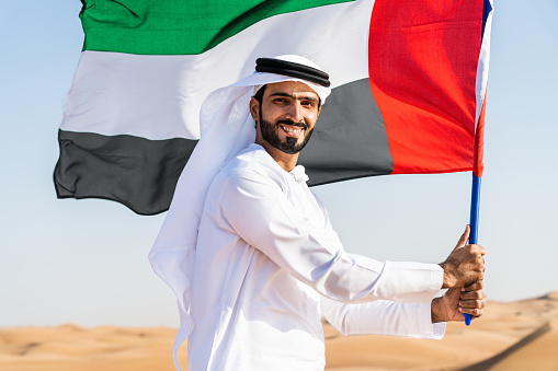 Middle-eastern man wearing traditional emirati arab kandura  in the desert holding the UAE flag - Arabian muslim adult person at the sand dunes in Dubai celebrating patriotism on national day