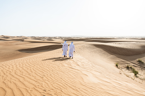 Two middle-eastern men wearing traditional emirati arab kandura bonding in the desert - Arabian muslim friends meeting at the sand dunes in Dubai