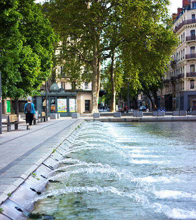 Lyon, France: A woman walks by the fountain on Place de la Republique. The water shoots into a large shallow pond, shaded by huge plane trees. This area is part of the UNESCO World Heritage site designation.