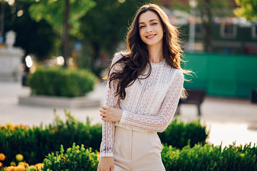 Fashion photo of a happy young brunette posing on a city square downtown and smiling at the camera. A boho girl is standing in a city park and making eye contact.