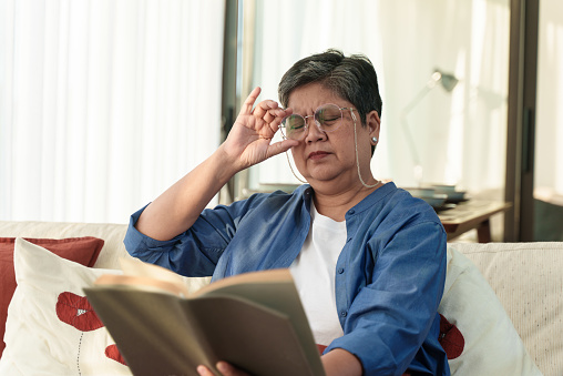 Portrait of Senior Asian woman with farsightedness presbyopia, taking off eyeglasses while reading book, poor vision, Long sighted problems. Senior health condition problem.