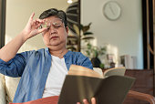 Portrait of Senior Asian woman with farsightedness presbyopia, taking off eyeglasses while reading book, poor vision, Long sighted problems. Senior health condition problem.