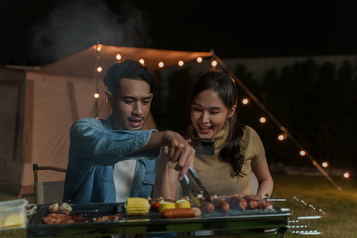 Young Asian couple grilling pork barbecue steak on charcoal stove at night during outdoor camping holiday travel in nature. Outdoor cooking camping travel concept.