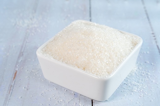 A white ceramic bowl containing granulated white sugar sits on a pristine white tablecloth, ready to be used for recipes