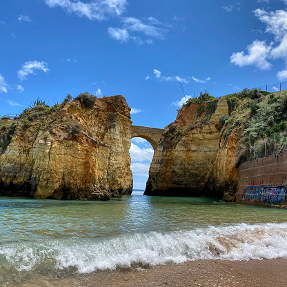 View of Roman bridge with a view of the beach. Water waves crashing on the beach. View of the sky in the arch of the bridge. No people in the photo.