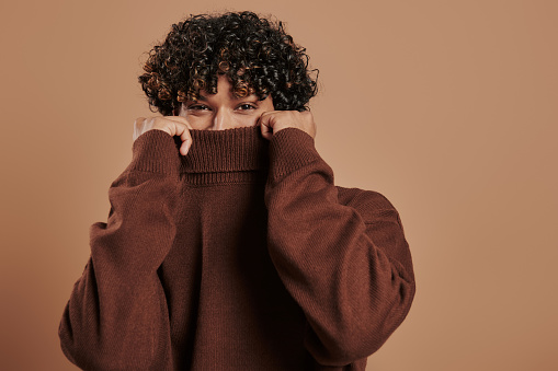 Playful young man hiding half face behind sweater collar against brown background