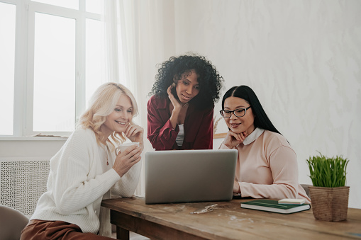 Three confident mature women using laptop and smiling while sitting at the desk together