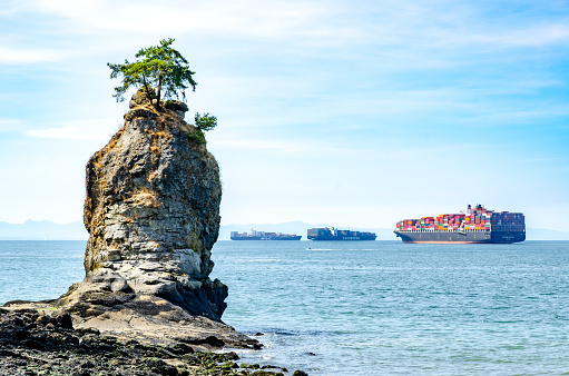 Vancouver, British Columbia, Canada â August 10, 2021: Siwash Rock in Stanley Park, Vancouver, BC and three commercial ships anchored in the background