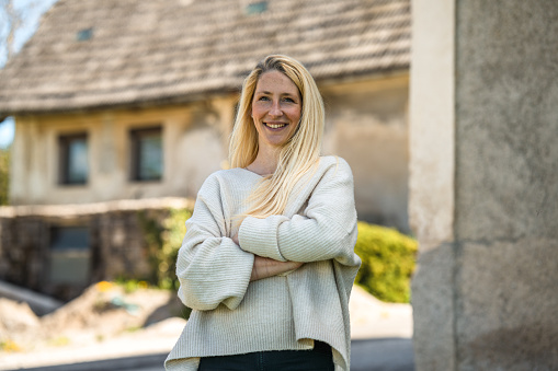 Portrait of a cheerful Caucasian female standing outdoors with arms crossed. Old house in the background.