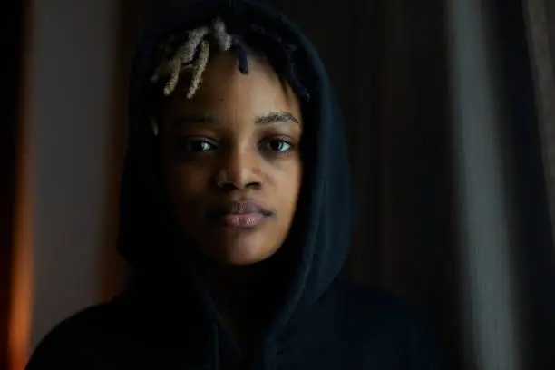 An African descent lady with black hoodie looking near window in her dark color room