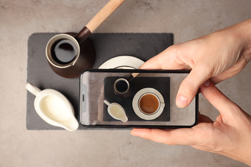 Woman taking photo of Turkish coffee and milk with smartphone at grey table, top view