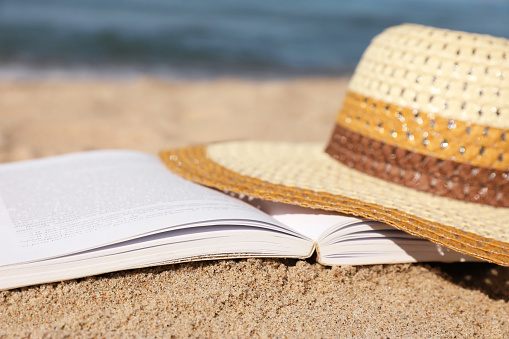 Open book and hat on sandy beach near sea, closeup