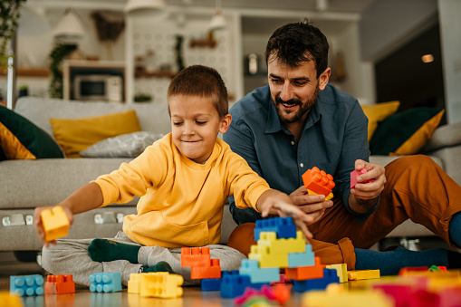 A caring young single father helps cute kid son play on a warm floor together, a happy family dad and little child boy having fun building a constructor tower from colorful blocks.