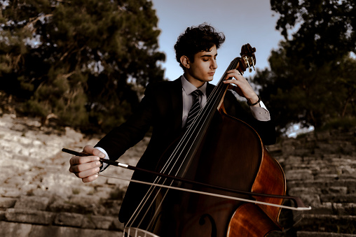 Young musician in a black suit plays double bass in the historical arena and in nature. He sits on a rock and makes music in the summer. Turkey, Antalya, Phaselis ancient city.