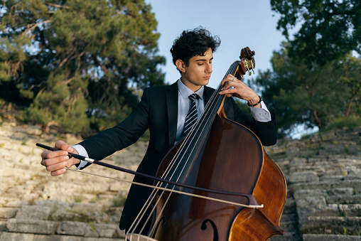 Young musician in a black suit plays double bass in the historical arena and in nature. He sits on a rock and makes music in the summer. Turkey, Antalya, Phaselis ancient city.