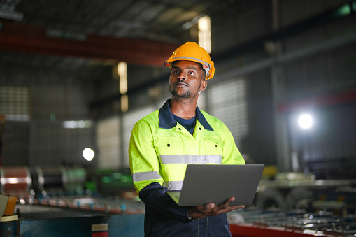 Factory engineer African man standing confidence with laptop. Worker works at heavy machine at industry factory. with machinery equipment plant technology.