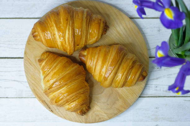 Fresh baked croissants on wooden cutting board on white wooden table with flowers Irises. Flat lay.