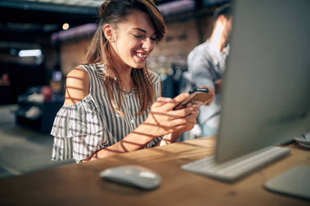 istock Beautiful young woman sitting at table in front of desktop computer using smartphone, feeling joyful. 1488107677