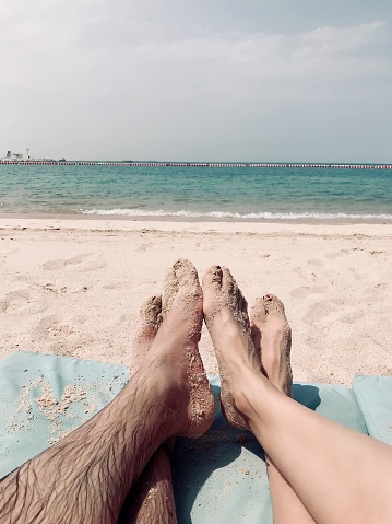 Stock photo showing elevated view of three unrecognisable people standing barefoot on compact wet sand as tide rolls in. An Asian man stood in the middle of a caucasian man and a white woman. Racial equality concept