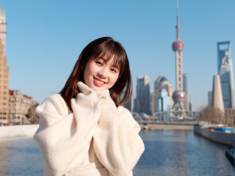 Beautiful young woman with black long hair in white skirt smiling with Shanghai city bund landmarks background in sunny day. Emotions, people, beauty, travel and lifestyle concept.