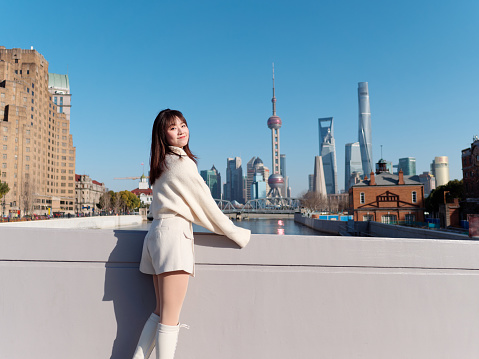 Beautiful young woman with black long hair in white skirt smiling with Shanghai city bund landmarks background in sunny day. Emotions, people, beauty, travel and lifestyle concept.