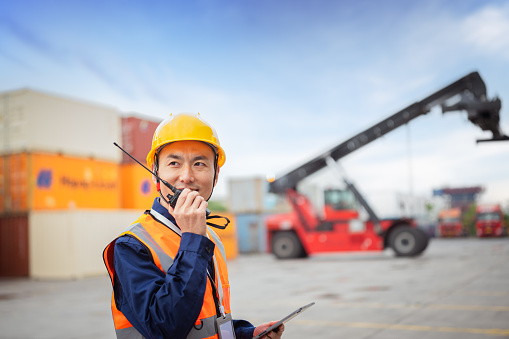 The Asian male management and transportation control engineer stands in front of the container and communicates with each other through walkie talkie. Conduct outdoor operations and control shipping at the loading dock.